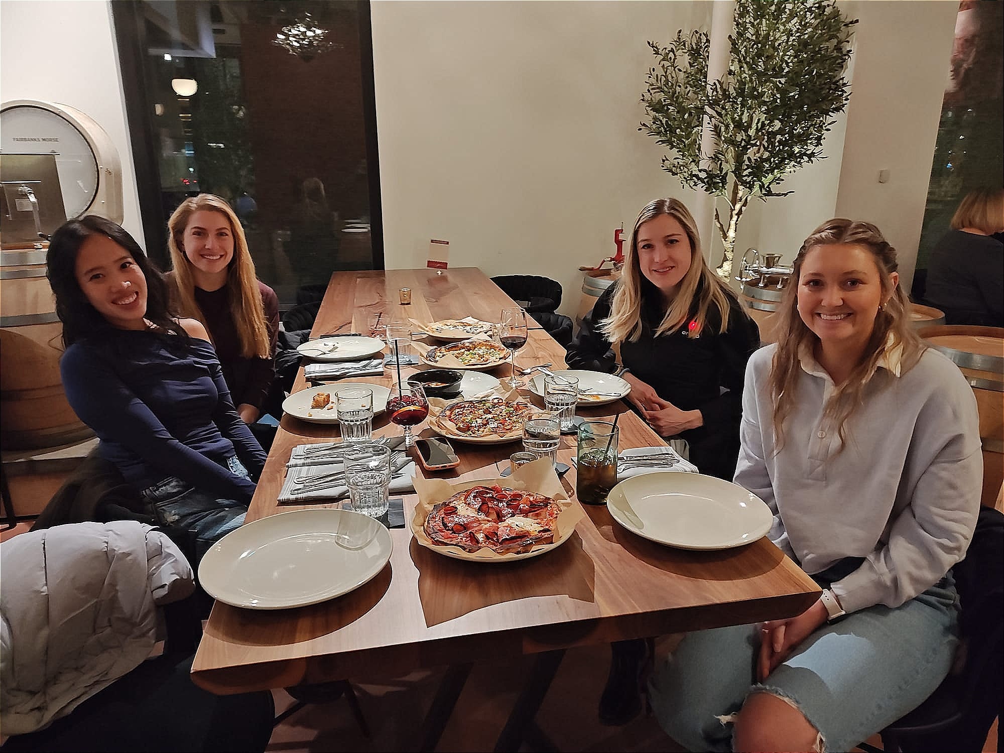 four women sitting at a dining table with four pizzas