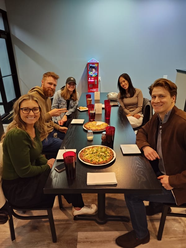 three women and two men smiling and sitting at a dining table with two pizzas